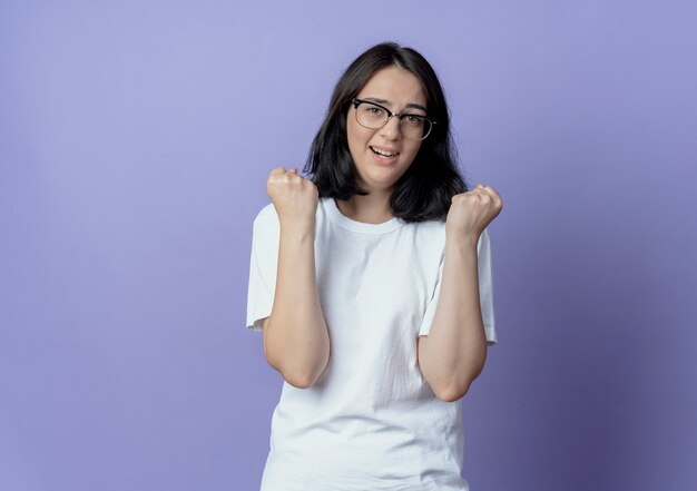 Impressionné jeune jolie fille caucasienne portant des lunettes serrant les poings isolés sur fond violet avec espace copie