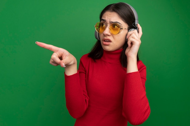Impressionné jeune jolie fille caucasienne portant des lunettes de soleil et des écouteurs saisissant des écouteurs regardant et pointant sur le côté isolé sur un mur vert