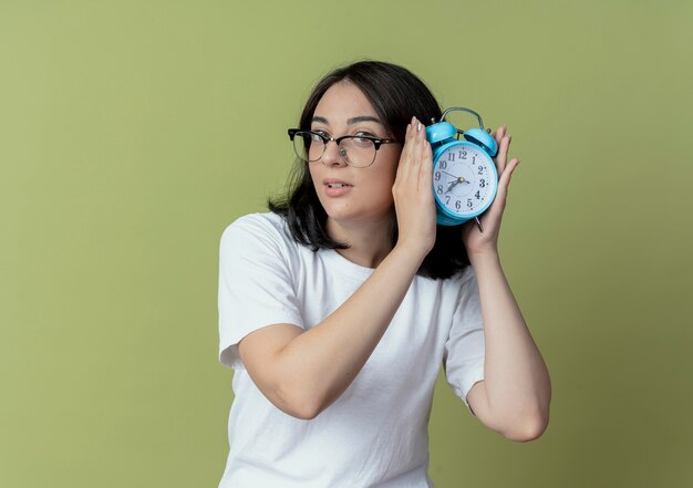 Impressionné jeune jolie fille caucasienne portant des lunettes tenant un réveil isolé sur fond vert olive avec espace copie