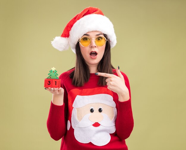 Impressionné jeune jolie fille caucasienne portant pull du père Noël et bandeau avec des lunettes tenant et pointant sur le jouet de l'arbre de Noël avec date