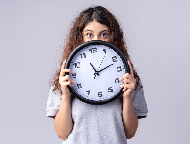 Impressionné jeune jolie fille caucasienne tenant une horloge par derrière isolé sur un mur blanc avec espace de copie