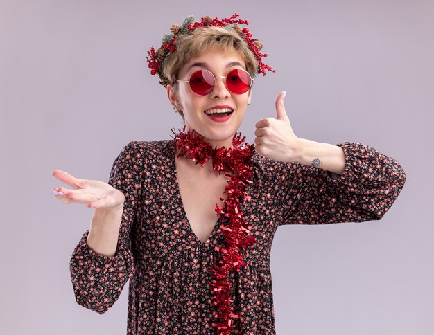 Impressionné jeune jolie fille portant couronne de tête de Noël et guirlande de guirlandes autour du cou avec des lunettes regardant la caméra montrant la main vide et le pouce vers le haut isolé sur fond blanc