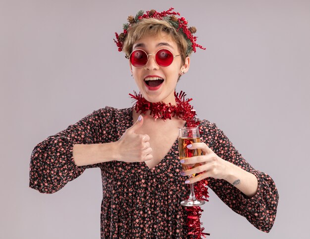 Impressionné jeune jolie fille portant couronne de tête de Noël et guirlande de guirlandes autour du cou avec des verres tenant un verre de champagne regardant la caméra montrant le pouce vers le haut isolé sur fond blanc