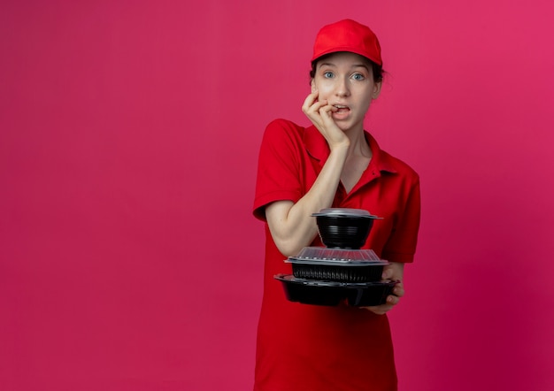 Impressionné Jeune Jolie Livreuse Portant Un Uniforme Rouge Et Une Casquette Tenant Des Contenants De Nourriture Et Se Mordant Les Doigts Isolés Sur Fond Cramoisi Avec Espace De Copie