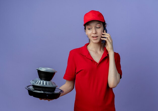 Impressionné Jeune Jolie Livreuse Portant Un Uniforme Rouge Et Une Casquette Tenant Et Regardant Les Contenants De Nourriture Et Parler Au Téléphone Isolé Sur Fond Violet Avec Espace De Copie