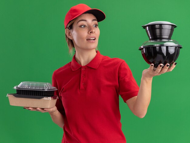 Impressionné jeune jolie livreuse en uniforme détient des emballages alimentaires et se penche sur les récipients alimentaires sur vert