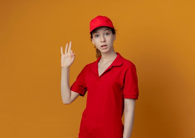 Impressionné jeune jolie livreuse en uniforme rouge et cap faisant signe ok regardant la caméra isolée sur fond orange avec copie espace