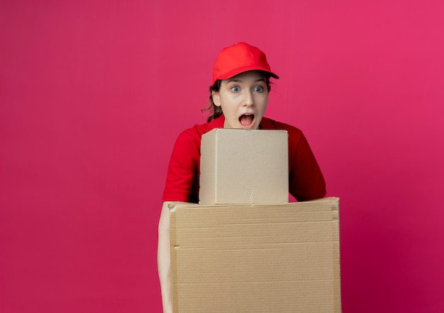 Photo gratuite impressionné jeune jolie livreuse en uniforme rouge et cap tenant des boîtes en carton à tout droit isolé sur fond cramoisi avec espace de copie