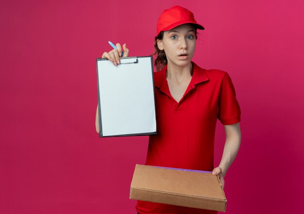 Impressionné jeune jolie livreuse en uniforme rouge et cap tenant le stylo paquet de pizza et montrant le presse-papiers isolé sur fond cramoisi avec espace de copie