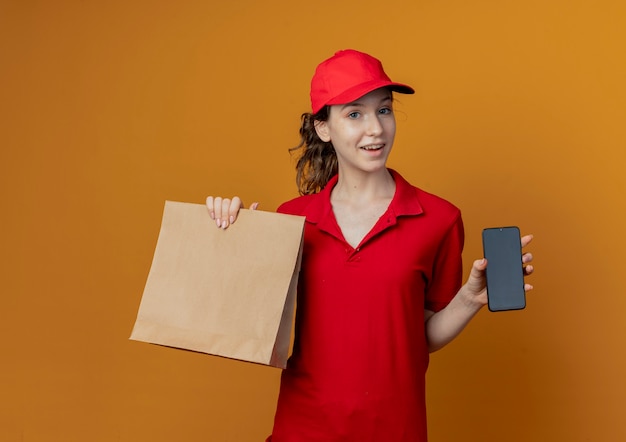 Impressionné jeune jolie livreuse en uniforme rouge et chapeau montrant le paquet de papier et téléphone mobile isolé sur fond orange avec copie espace