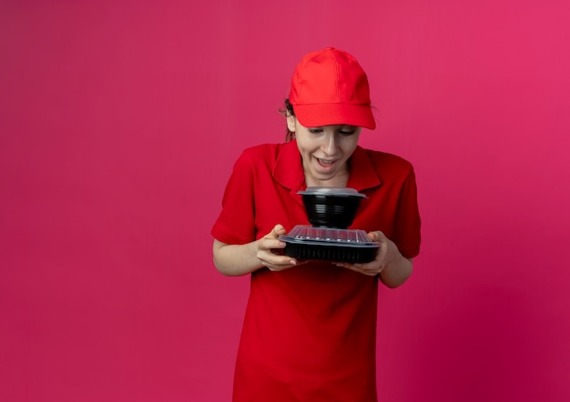 Impressionné jeune jolie livreuse vêtue d'un uniforme rouge et d'une casquette tenant et regardant des contenants de nourriture