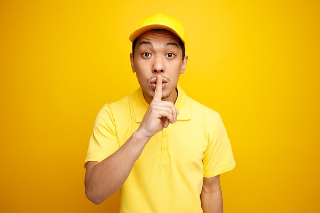 Impressionné jeune livreur portant casquette et uniforme faisant le geste de silence