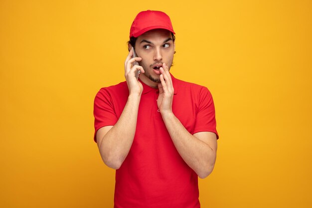 impressionné jeune livreur portant l'uniforme et la casquette regardant le côté en gardant la main près de la bouche chuchotant au téléphone isolé sur fond jaune