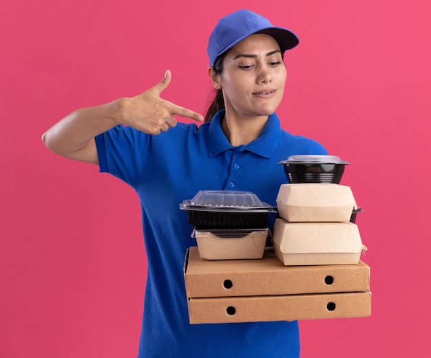 Impressionné jeune livreuse portant l'uniforme avec cap holding et points à des contenants de nourriture sur des boîtes de pizza isolé sur mur rose