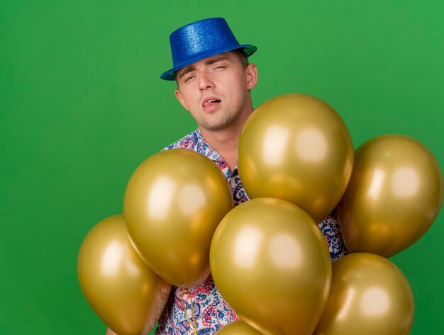 Impressionné jeune mec portant chapeau bleu debout parmi les ballons isolés sur vert