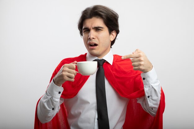 Impressionné jeune mec de super-héros portant une cravate tenant et des points à une tasse de café isolé sur fond blanc