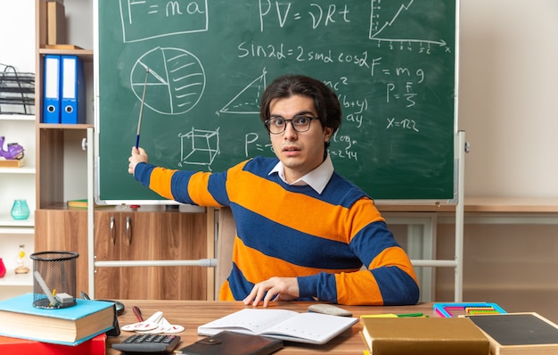 impressionné jeune professeur de géométrie caucasien portant des lunettes assis au bureau avec des fournitures scolaires en classe gardant la main sur le bureau regardant l'avant pointant avec un bâton de pointeur au tableau