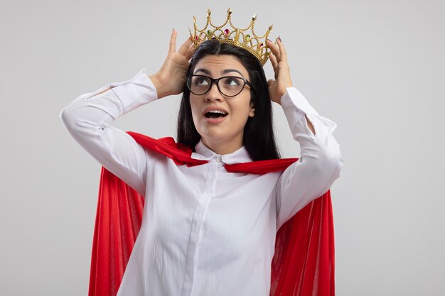 Impressionné jeune superwoman portant des lunettes et couronne touchant la couronne en levant isolé sur mur blanc