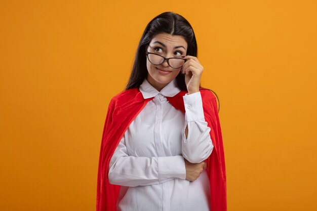 Impressionné jeune superwoman portant des lunettes en les saisissant à côté isolé sur mur orange