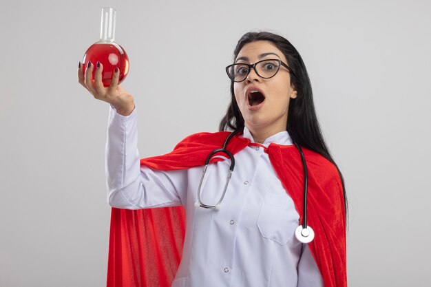 Impressionné jeune superwoman portant des lunettes et un stéthoscope soulevant un ballon chimique avec un liquide rouge jusqu'à l'avant isolé sur un mur blanc