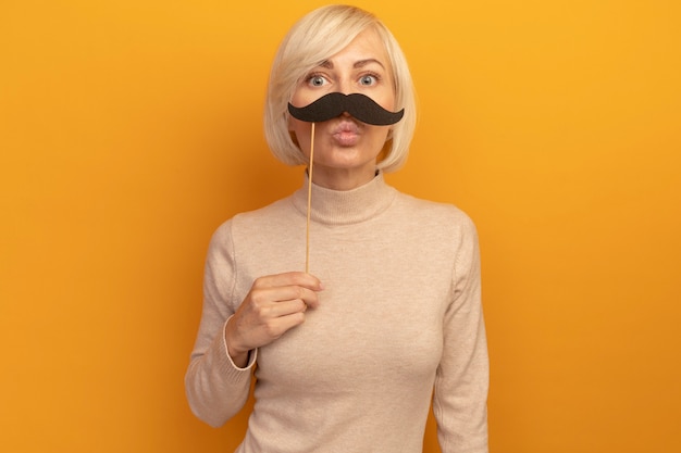 Impressionné jolie blonde femme slave tient une fausse moustache sur bâton isolé sur mur orange