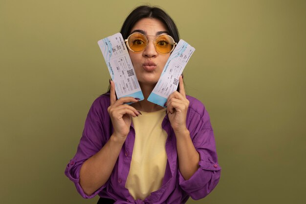 Impressionné jolie femme brune à lunettes de soleil détient des billets d'avion près du visage isolé sur mur vert olive