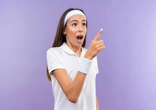 Impressionné jolie fille sportive portant un bandeau et un bracelet pointant sur le côté isolé sur un mur violet