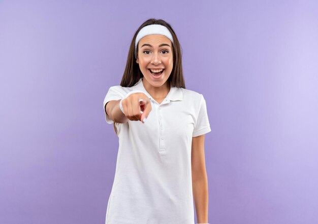 Impressionné jolie fille sportive portant un bandeau et un bracelet pointant isolé sur un mur violet