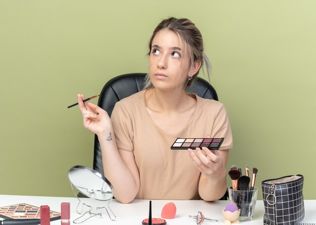 Impressionné en levant la belle jeune fille assise à table avec des outils de maquillage tenant une brosse avec une palette de fards à paupières isolée sur un mur vert olive