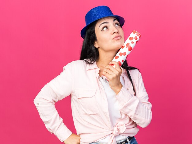 Impressionné en levant la belle jeune fille portant un chapeau de fête tenant un canon à confettis mettant la main sur la hanche