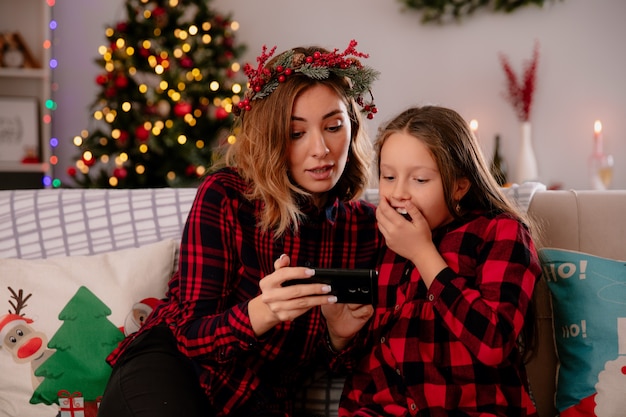 Impressionné mère et fille regardant quelque chose sur le téléphone assis sur un canapé et profiter du temps de Noël à la maison