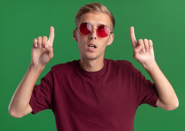 Impressionné à la recherche d'un jeune beau mec portant une chemise rouge et des lunettes à point isolé sur mur vert