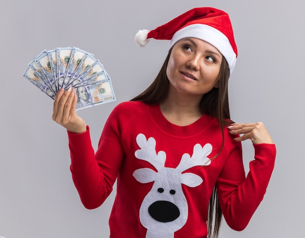 Impressionné à la recherche de jeune fille asiatique portant chapeau de Noël avec chandail tenant de l'argent mettant la main sur l'épaule isolé sur fond blanc