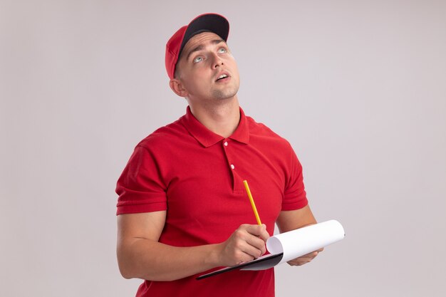 Impressionné à la recherche de jeune livreur en uniforme avec capuchon tenant le presse-papiers avec un crayon isolé sur un mur blanc