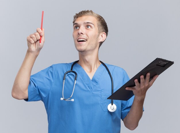 Impressionné à la recherche d'un jeune médecin de sexe masculin portant l'uniforme de médecin avec stéthoscope tenant le presse-papiers avec un crayon isolé sur un mur blanc