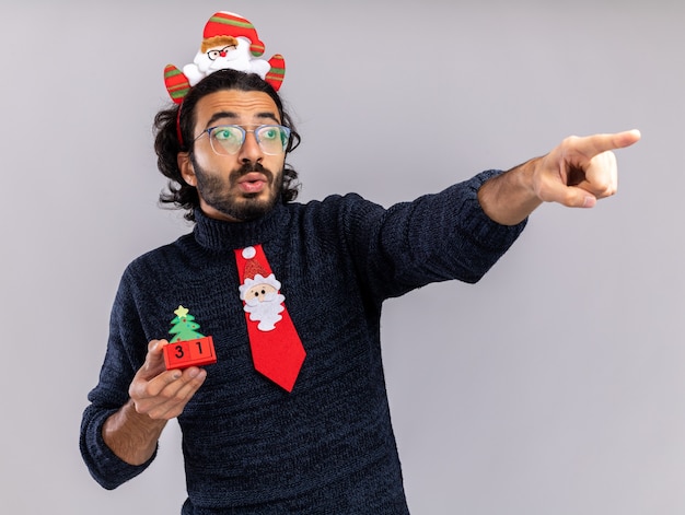 Photo gratuite impressionné regardant côté jeune beau mec portant une cravate de noël avec cerceau de cheveux tenant des points de jouets de noël à côté isolé sur fond blanc