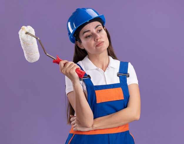 Impressionné en regardant côté jeune femme constructeur en uniforme tenant une brosse à rouleau isolé sur un mur violet