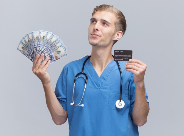 Impressionné regardant côté jeune médecin de sexe masculin portant l'uniforme de médecin avec stéthoscope tenant de l'argent et une carte de crédit isolé sur un mur blanc