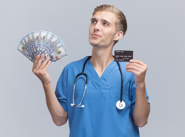 Impressionné regardant côté jeune médecin de sexe masculin portant l'uniforme de médecin avec stéthoscope tenant de l'argent et une carte de crédit isolé sur un mur blanc