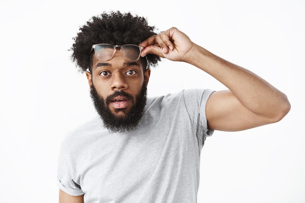 Impressionné et surpris, beau mec afro-américain enlevant des lunettes comme étant charmé par la beauté tenant des lunettes sur le front bouche ouverte d'étonnement en regardant la caméra sur un mur gris