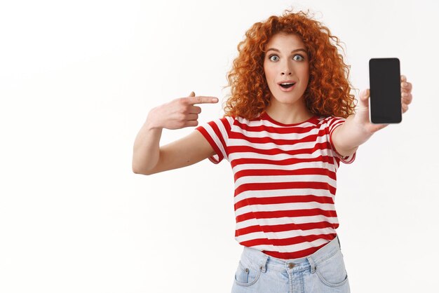 Impressionné surpris rousse fasciné jeune femme curlyhaired gingembre fille pointant l'écran du smartphone étonné génial nouvelle application recommander application cool debout fond blanc