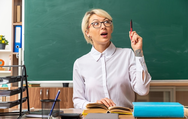 impressionnée jeune enseignante blonde portant des lunettes assis au bureau avec des outils scolaires en classe pointant vers le haut avec un stylo en gardant la main sur un livre ouvert regardant de côté