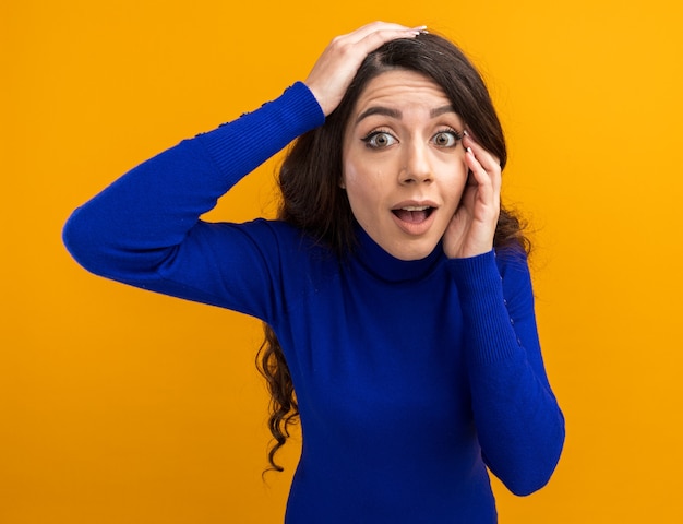 impressionnée jeune jolie femme touchant le visage en gardant la main sur la tête en regardant l'avant isolé sur le mur orange