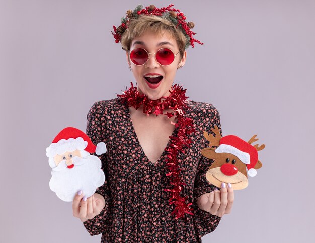 impressionnée jeune jolie fille portant une couronne de tête de noël et une guirlande de guirlandes autour du cou avec des lunettes tenant des ornements de papier de renne de noël et de père noël isolés sur un mur blanc