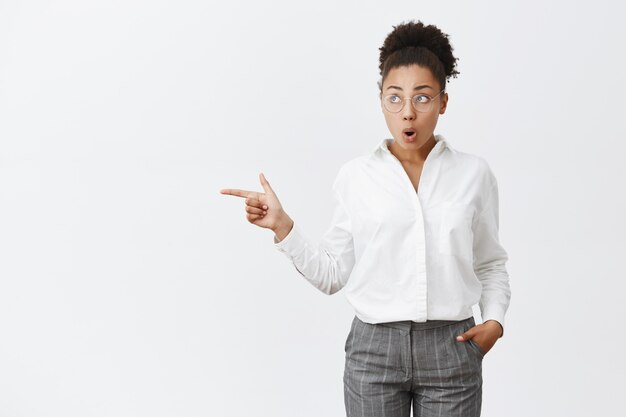 impressionnée et surprise jolie femme à la peau foncée avec des cheveux bouclés dans des lunettes et une chemise à col blanc, tenant la main dans la poche, pliant les lèvres par curiosité, regardant et pointant vers la gauche