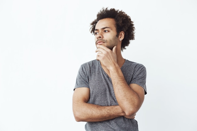 Incertain homme africain pensant regarder de côté sur le mur blanc, décider d'acheter des gadgets en ligne ou un étudiant essayant de se souvenir d'une fête.