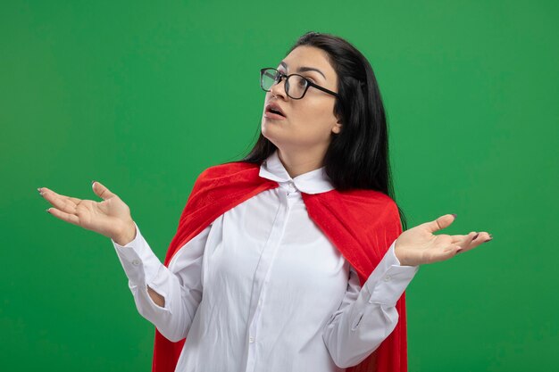 Incompréhension confuse jeune superwoman portant des lunettes tenant les mains vides et ne comprends pas ce qui se passe à droite isolé sur mur vert