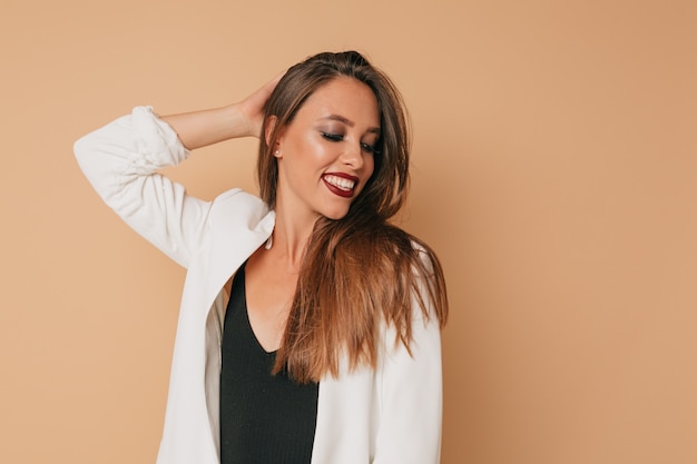 Incroyable élégante femme souriante avec rouge à lèvres vigne portant une veste blanche posant sur un mur beige, préparation pour la fête, mur isolé