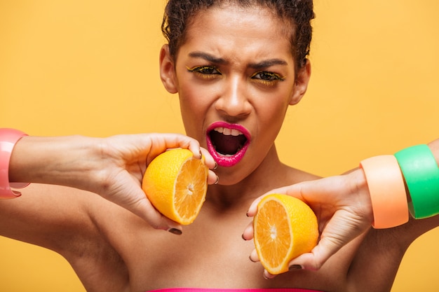 Incroyable femme afro-américaine conceptuelle mettant deux parties d'orange mûre de nouveau ensemble sur l'appareil photo isolé, sur le mur jaune