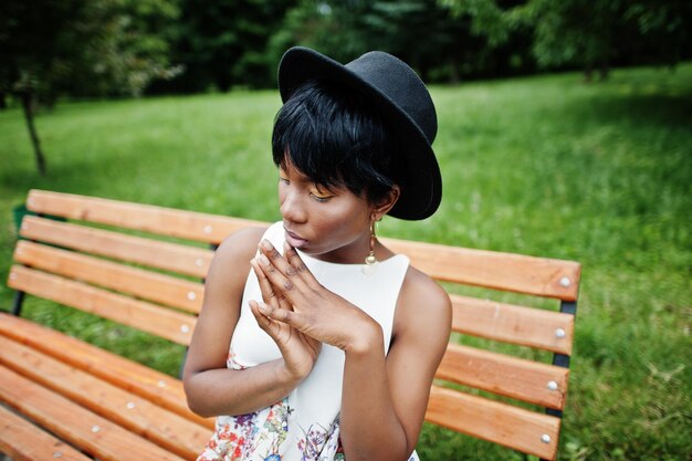 Incroyable femme modèle afro-américaine en pantalon vert et chapeau noir posé sur un banc au parc